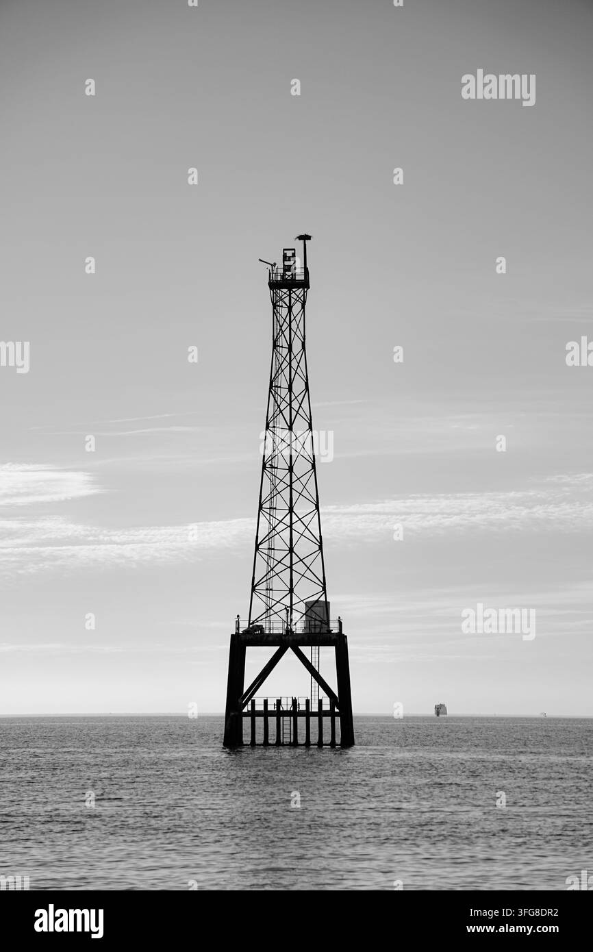 Ein hoher Metallturm steht allein im Ozean, erhebt sich über ruhiges Wasser unter einem weichen Himmel und schafft eine beeindruckende Szene einer isolierten Küstenstruktur. Stockfoto