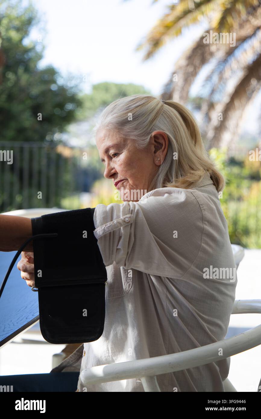 Seniorin mit hellem Hemd, die den Blutdruck mit einer schwarzen Armmanschette an einem runden Gartentisch überprüft Stockfoto