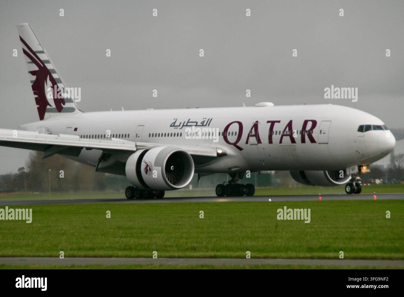 Ein Qatar Airways Boeing 777 Passagierflugzeug taxiert mit Umkehrschub auf einer nassen Landebahn unter bewölktem Himmel nach der Landung. Stockfoto