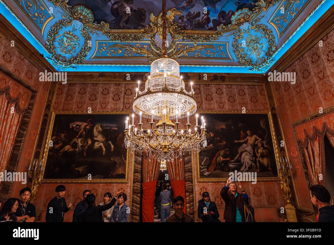 Madrid, Spanien: 7. Apr. 2025 – Eine Menschenmenge steht unter einem großen vergoldeten Kronleuchter in einem eleganten, mit Damast tapezierten Raum, an dessen Wänden Porträtgemälde hängen Stockfoto