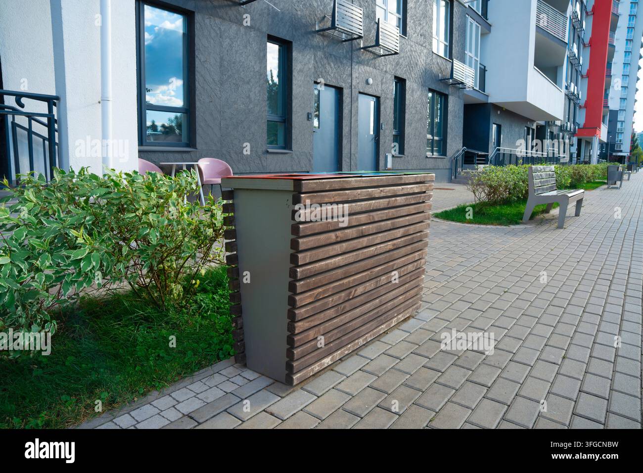 Moderner hölzerner Abfallbehälter auf einem gepflasterten Gehweg in der Nähe eines modernen Wohnapartmentgebäudes mit grüner Bepflanzung und einer Bank Stockfoto