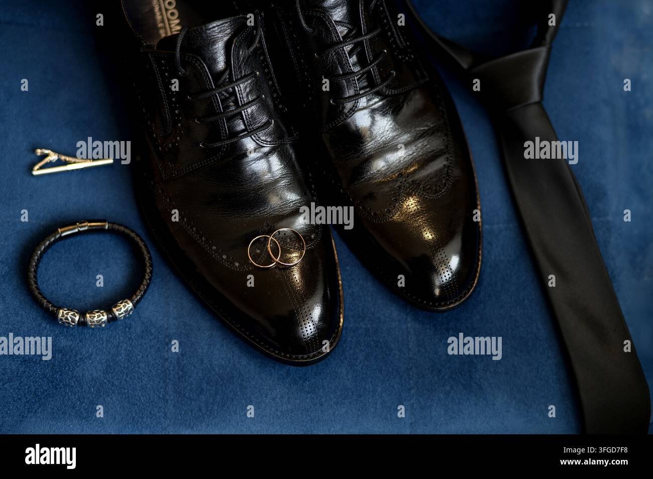 Elegante Bräutigam-Accessoires flach auf blauem Samtgrund, schwarze Lederschuhe im Oxford-Stil, Seidenkrawatte, goldene Eheringe und Lederarmband zur Schau gestellt Stockfoto