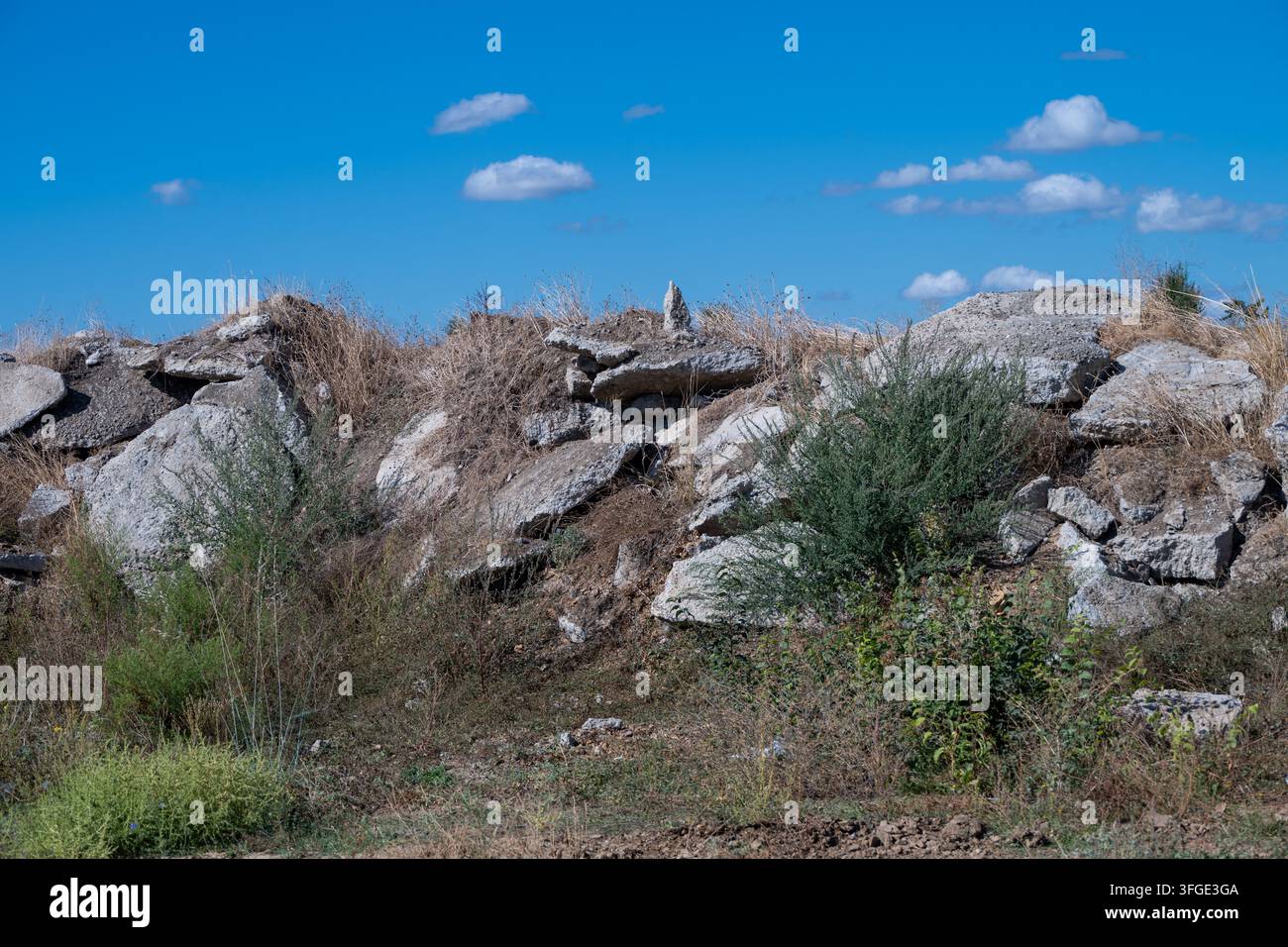 Ein Haufen abgelagerter Betonplatten und Schutt, überwuchert mit trockenem Gras und grünem Unkraut, liegt auf einem Damm unter einem strahlend blauen Himmel. Stockfoto