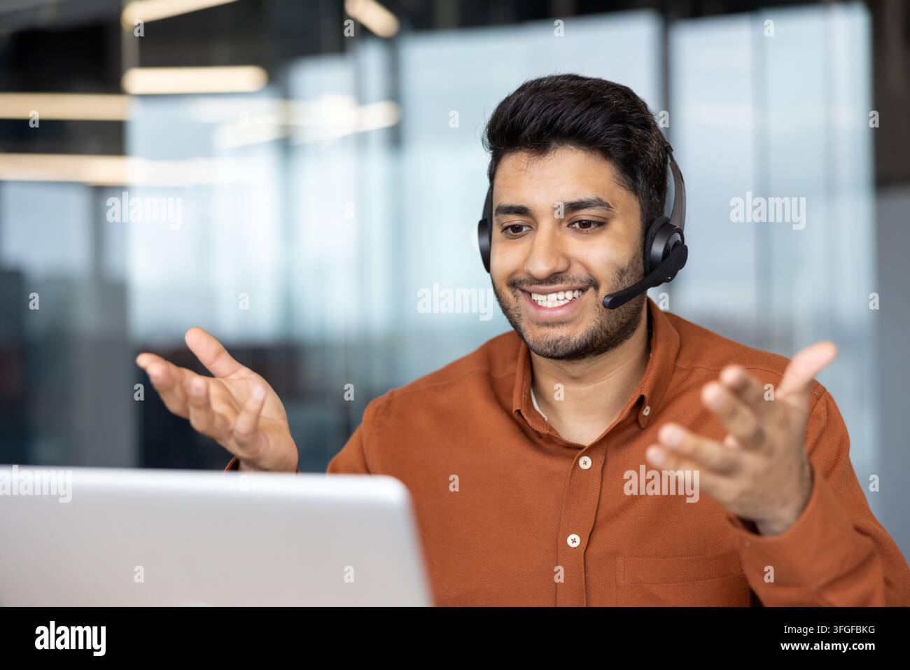 Lächelnder Mann mit Headset und orangefarbenem Hemd spricht während eines Online-Meetings von einem hellen Büroraum aus, verwendet einen Laptop und gestikuliert mit offenen Händen und zeigt freundliche, engagierte Kommunikation Stockfoto