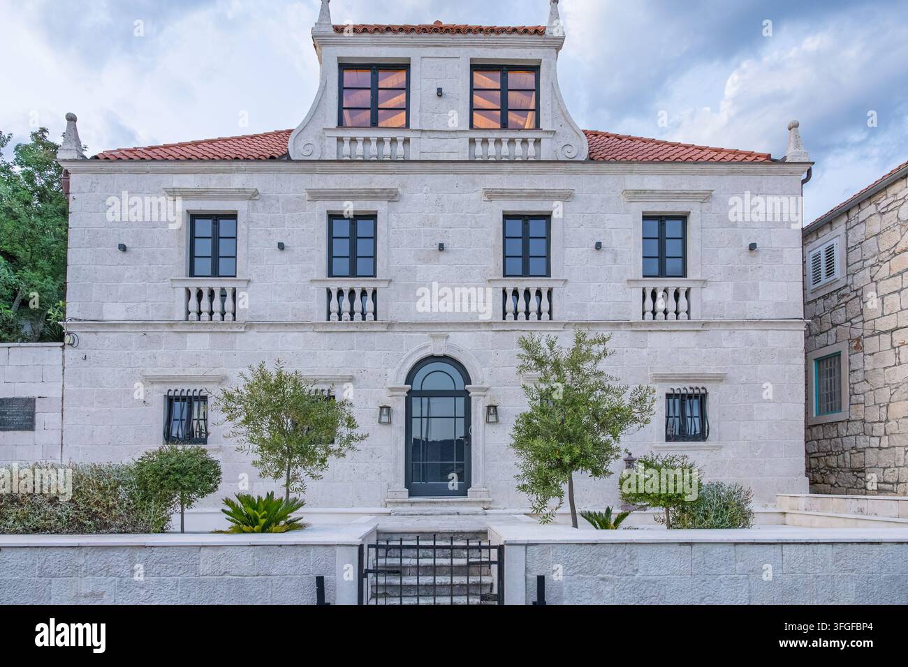 Eine symmetrische mediterrane Steinvillafassade mit schwarz gerahmten Fenstern, Terrakotta-Dachziegeln, dekorativen Balustraden und einem gewölbten Mittelrisalit Stockfoto