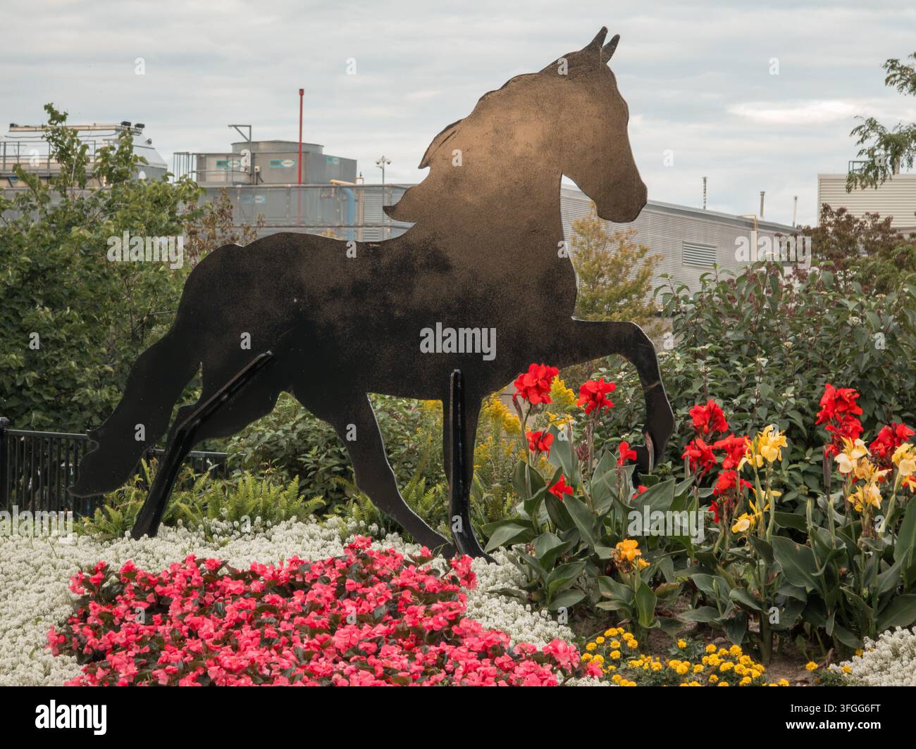 Eine auffällige Silhouette einer Pferdeskulptur steht inmitten lebendiger roter, gelber und rosa Blumen in einer üppigen Gartenumgebung. Diese Außenkunstinstallation - Smartphone-aufgenommenes Stockfoto