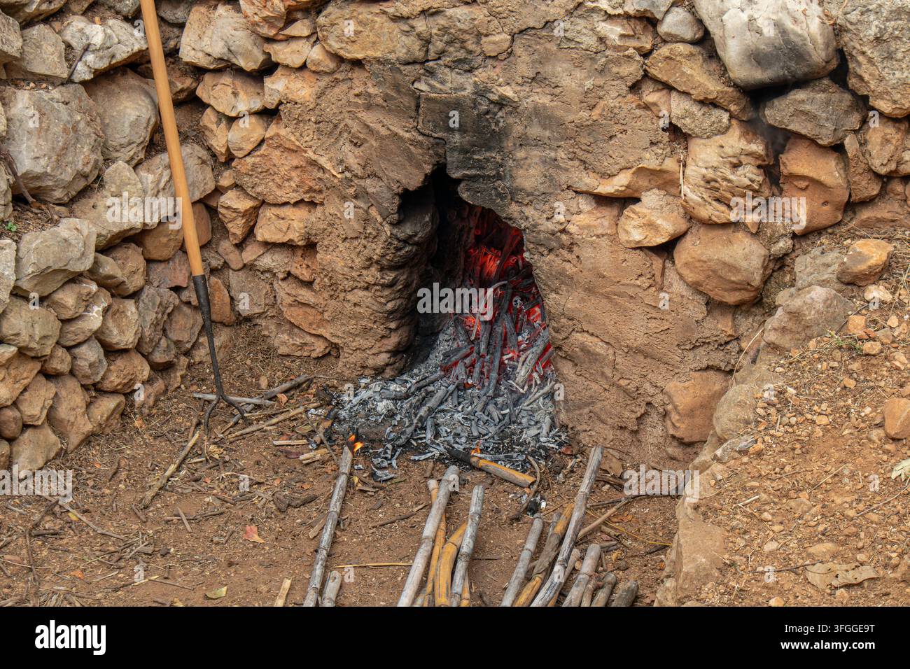 Der in eine Trockenmauer eingebaute Außensteinofen zeigt glühende rote Glut. In der Nähe liegen Stöcke auf dem Boden. Diese Szene spiegelt das Landleben wider, a Stockfoto