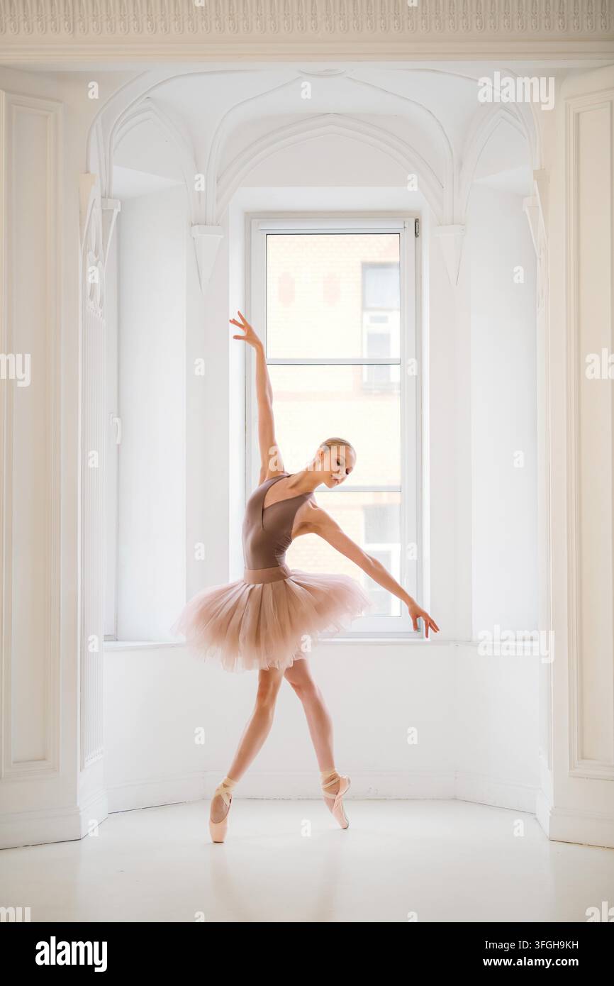 Ballerina in einem beigen Trikot und Tutu posiert anmutig auf dem Zehenspitzen in einem hellen Ballettstudio, ihre Reflexion im Spiegel vervollständigt die Szene. Stockfoto