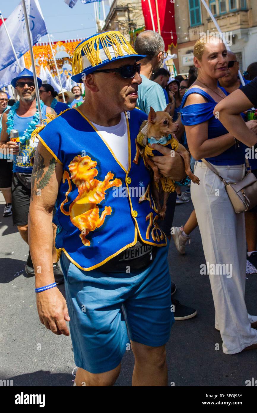 Zabbar, Malta - 12. September 2026, Mann in blauem Kostüm und Hut hält einen kleinen Hund bei einem farbenfrohen Straßenfestumzug Stockfoto