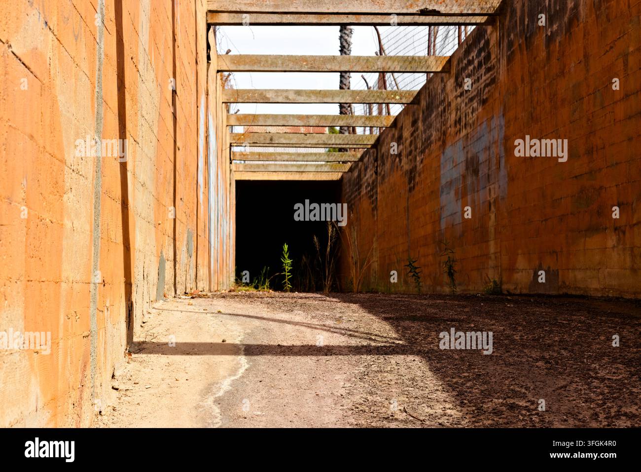 Verwitterte Betonwände und sich wiederholende Überkopfträger bilden einen schmalen trockenen Kanal mit tiefen Schatten und spärlichem Unkraut in Riverside, Kalifornien. Stockfoto