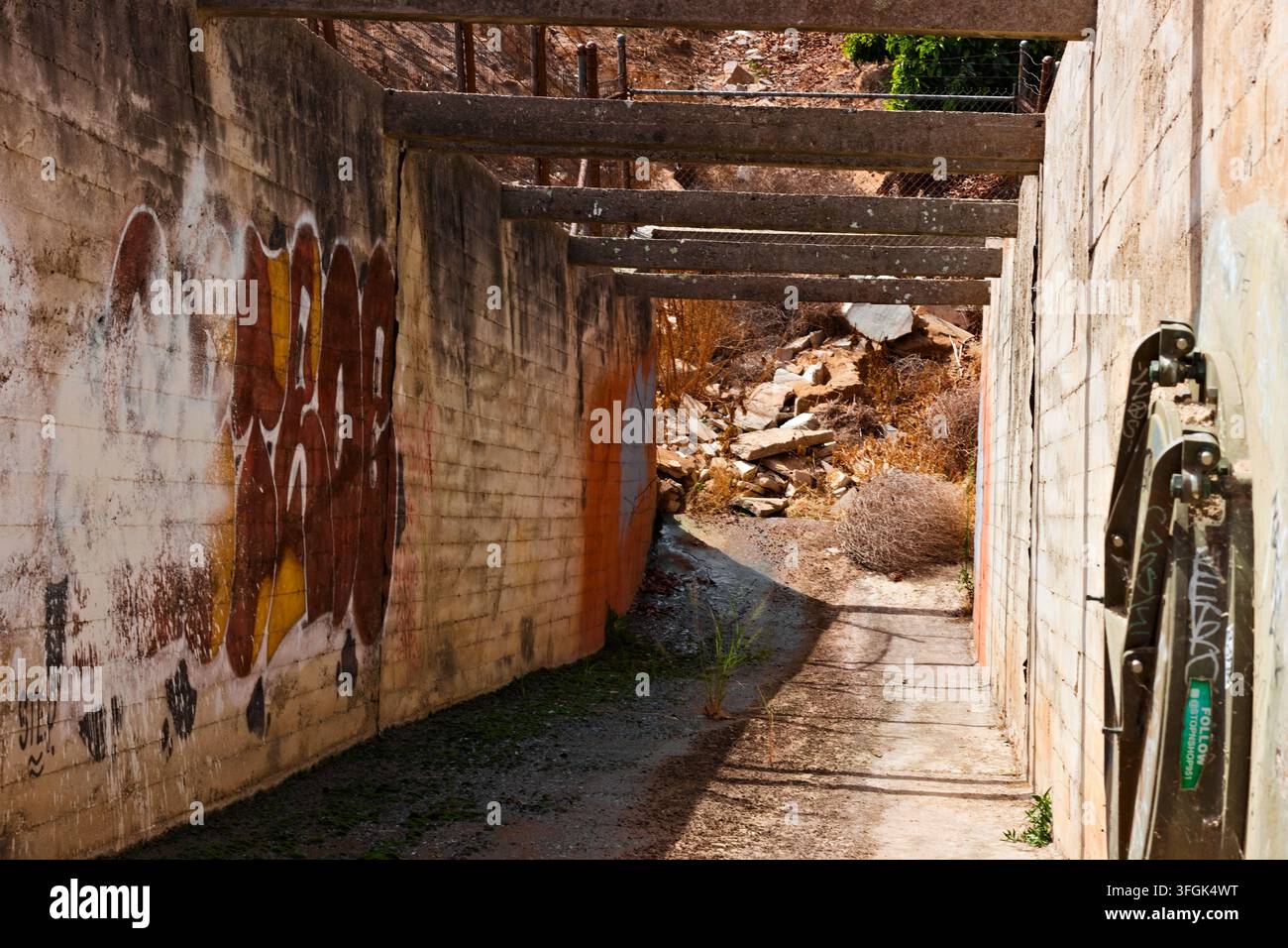 Verwitterte Betonwände und sich wiederholende Überkopfträger bilden einen schmalen trockenen Kanal mit tiefen Schatten und spärlichem Unkraut in Riverside, Kalifornien. Stockfoto