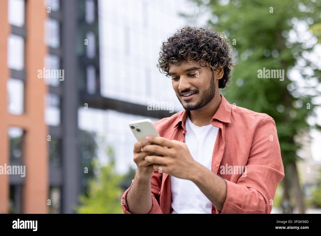 Lächelnder junger männlicher Universitätsstudent geht während der Pause auf dem Campusgelände und nutzt sein Smartphone. Stockfoto