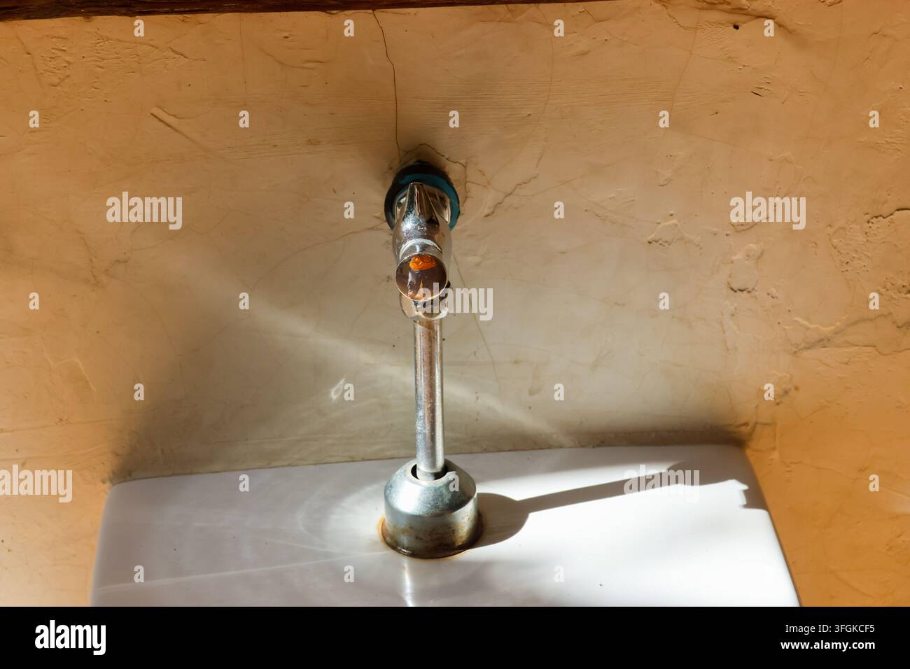 Ein Chrom-Wasserhahn, der an einer strukturierten Putzwand über einem weißen Waschbecken montiert ist, wird von warmem Sonnenlicht beleuchtet und wirft einen scharfen Schatten in einer rustikalen Inneneinrichtung Stockfoto