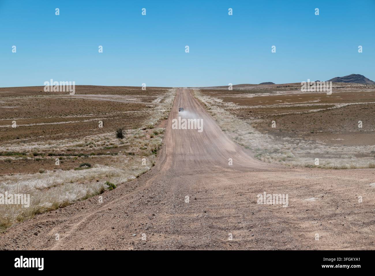 Weite Wüstenlandschaft in Namibia. Eine Schotterstraße erstreckt sich in die Ferne zu entfernten Bergen unter einem klaren blauen Himmel. Spärliche Vegetation ziert t Stockfoto