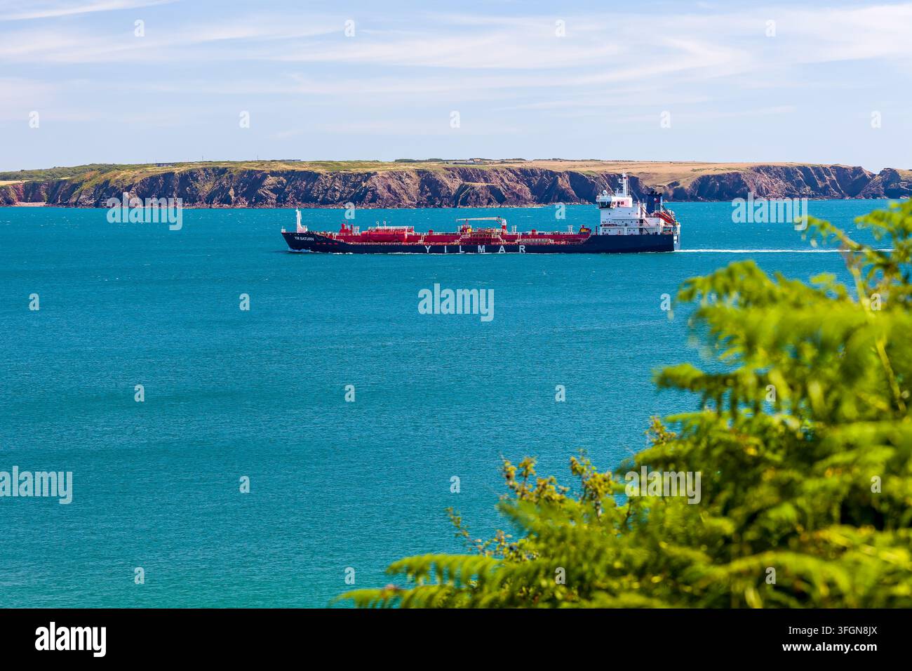 Handelstankerschiff, das auf offenem Wasser an schroffen Klippen vorbeifährt, Hochwinkelaufnahme eingerahmt von grünen Blättern, industrielle Seeroute in der Küstenestuarmündung Stockfoto