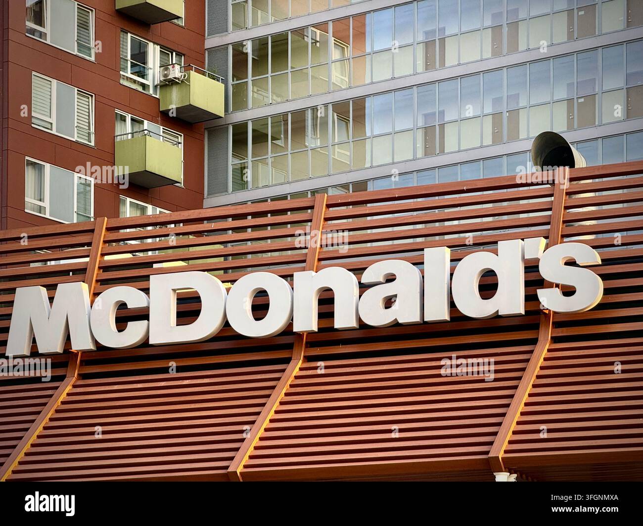 Nahaufnahme einer horizontalen Aufnahme eines weißen McDonald's-Schilds, das an einer modernen braunen Holzlattenfassade unter einem zeitgenössischen Glasgebäude montiert ist. - Smartphone-aufgenommenes Stockfoto
