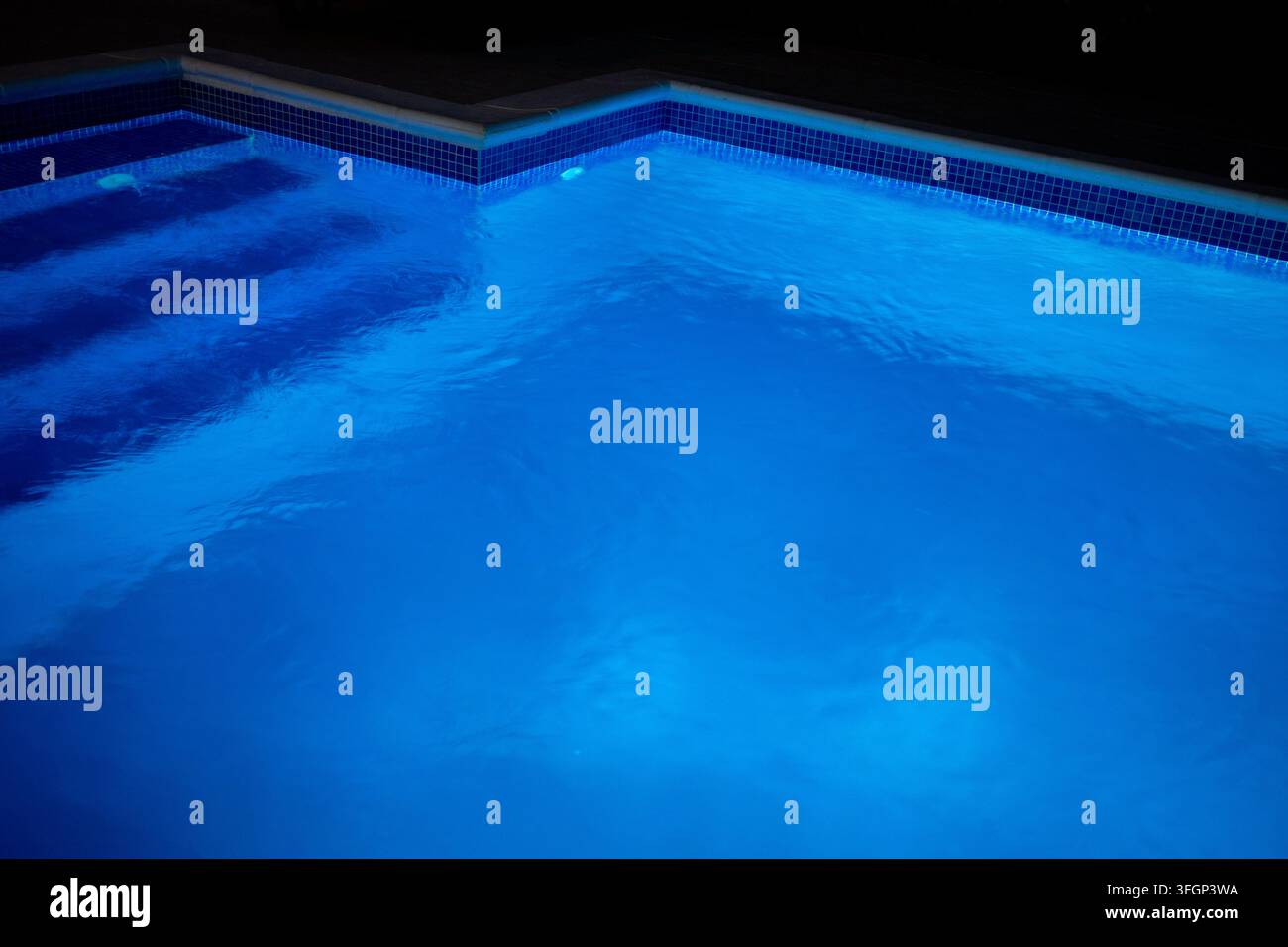 Menschen können ein Nachtschwimmen genießen, während blaue Lichter hell im Pool leuchten. Die Wasseroberfläche spiegelt das sanfte Glühen wider und zeigt die Stufen und das tiefe Ende. Stockfoto