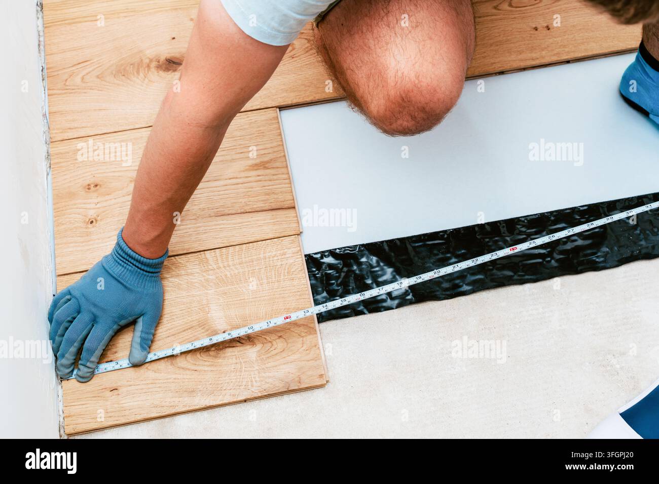 Bodenverlegung im Gange, während ein Arbeiter Holzplanken mit einem Maßband misst. Arbeiter richtet Bretter neben der Unterlage aus und zeigt präzise Renovierung Stockfoto