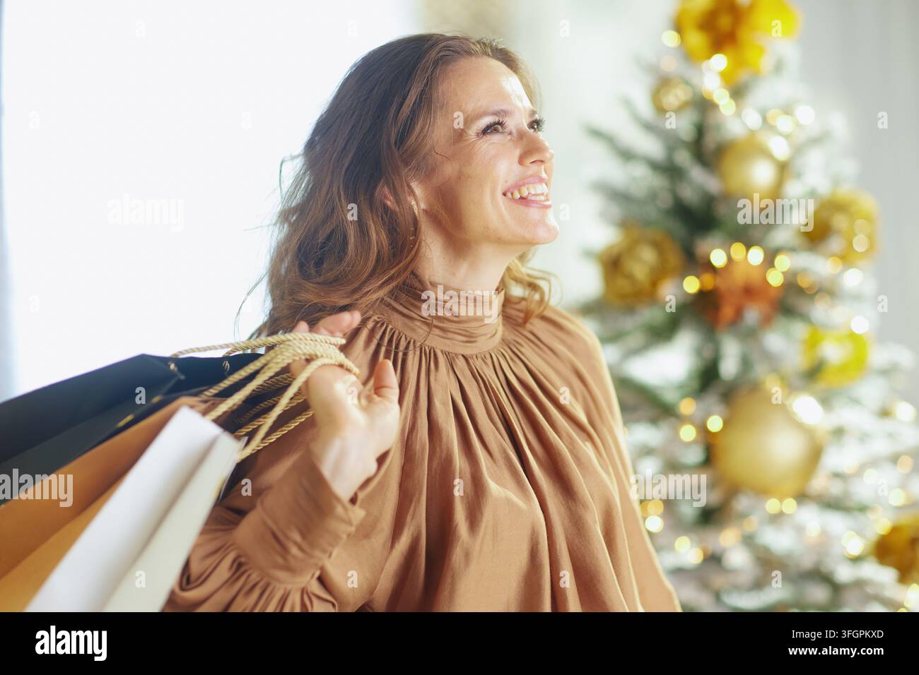 Strahlende Frau in goldener Satin-Rollobluse strahlt, trägt Einkaufstüten. Weich verschwommener Weihnachtsbaum mit goldenen Verzierungen und Lichterketten erzeugt hell Stockfoto