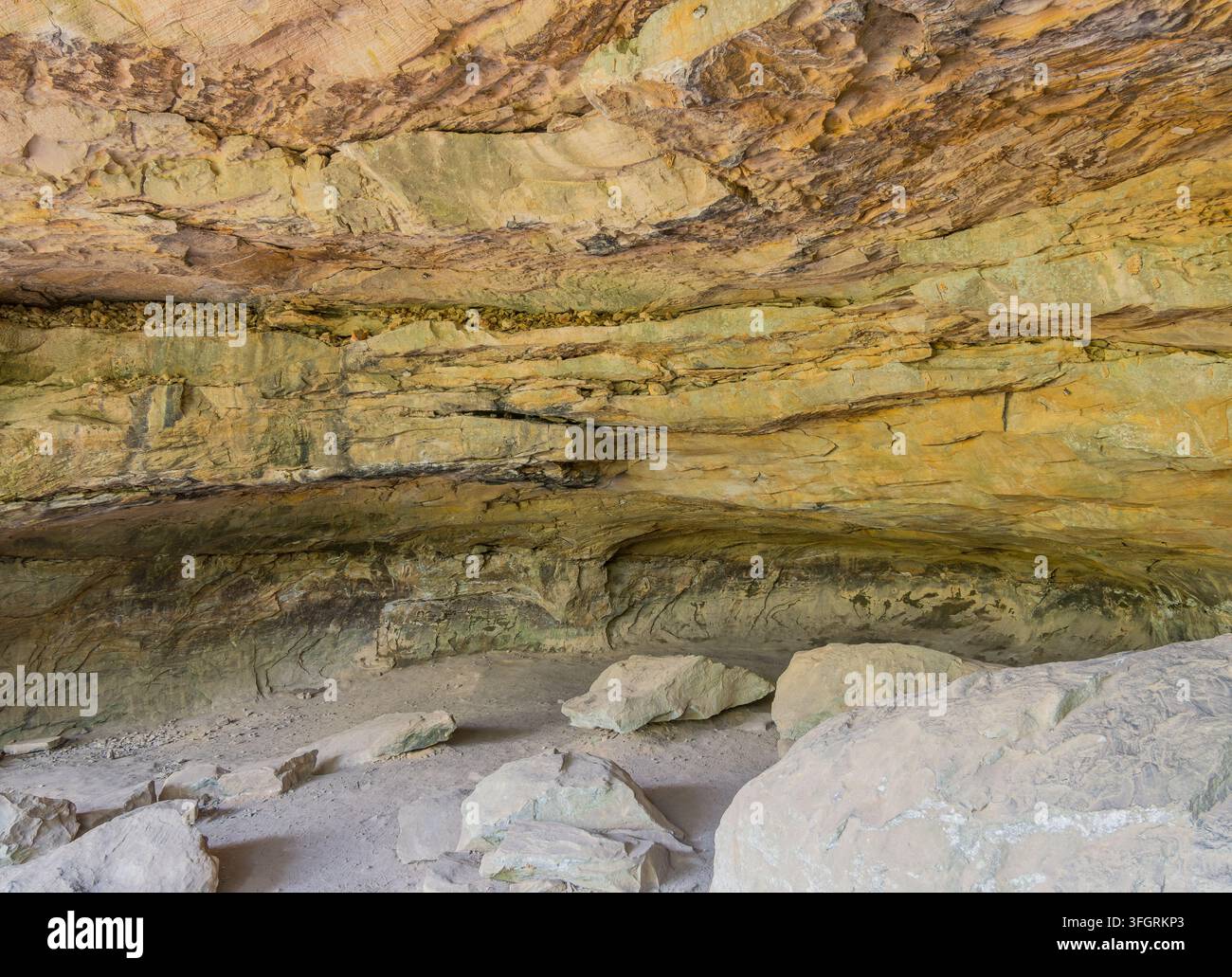 Sonnenlicht beleuchtet die strukturierte, geschichtete Sandsteindecke und die Wände eines natürlichen Felsenschutzes mit großen Felsbrocken, die auf dem sandigen Boden ruhen. Stockfoto