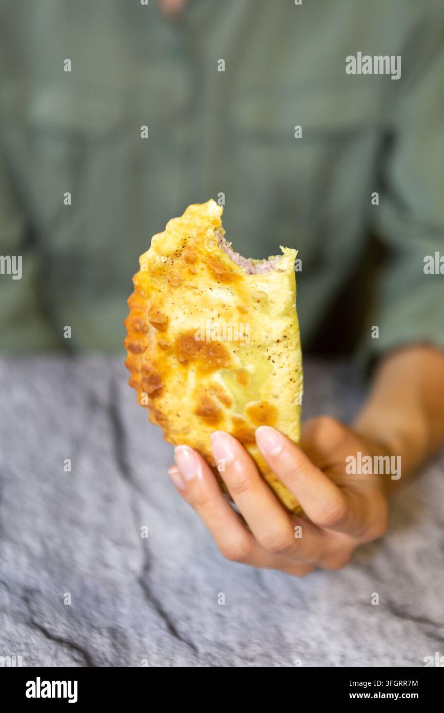Eine Person sitzt in einem Café und hält einen Cheburek in einer Hand. Der Teigtaschen haben eine goldene Kruste und sind mit Fleisch gefüllt. Die Mahlzeit findet drinnen statt Stockfoto