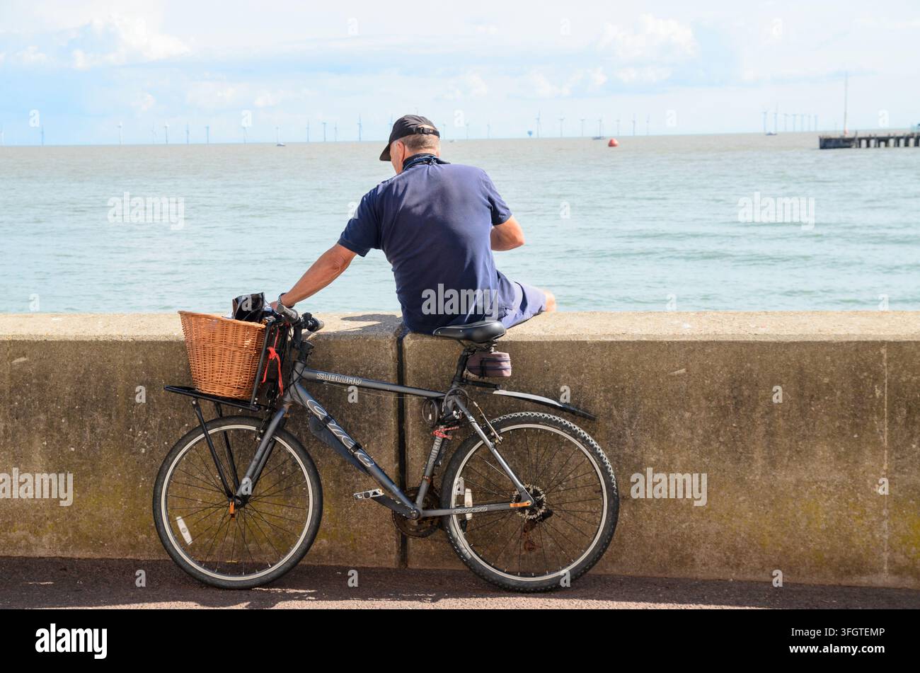 ein Mann, der auf einer Mauer sitzt, mit seinem Fahrrad, das an der Mauer lehnt, und aufs Meer blickt uk Stockfoto