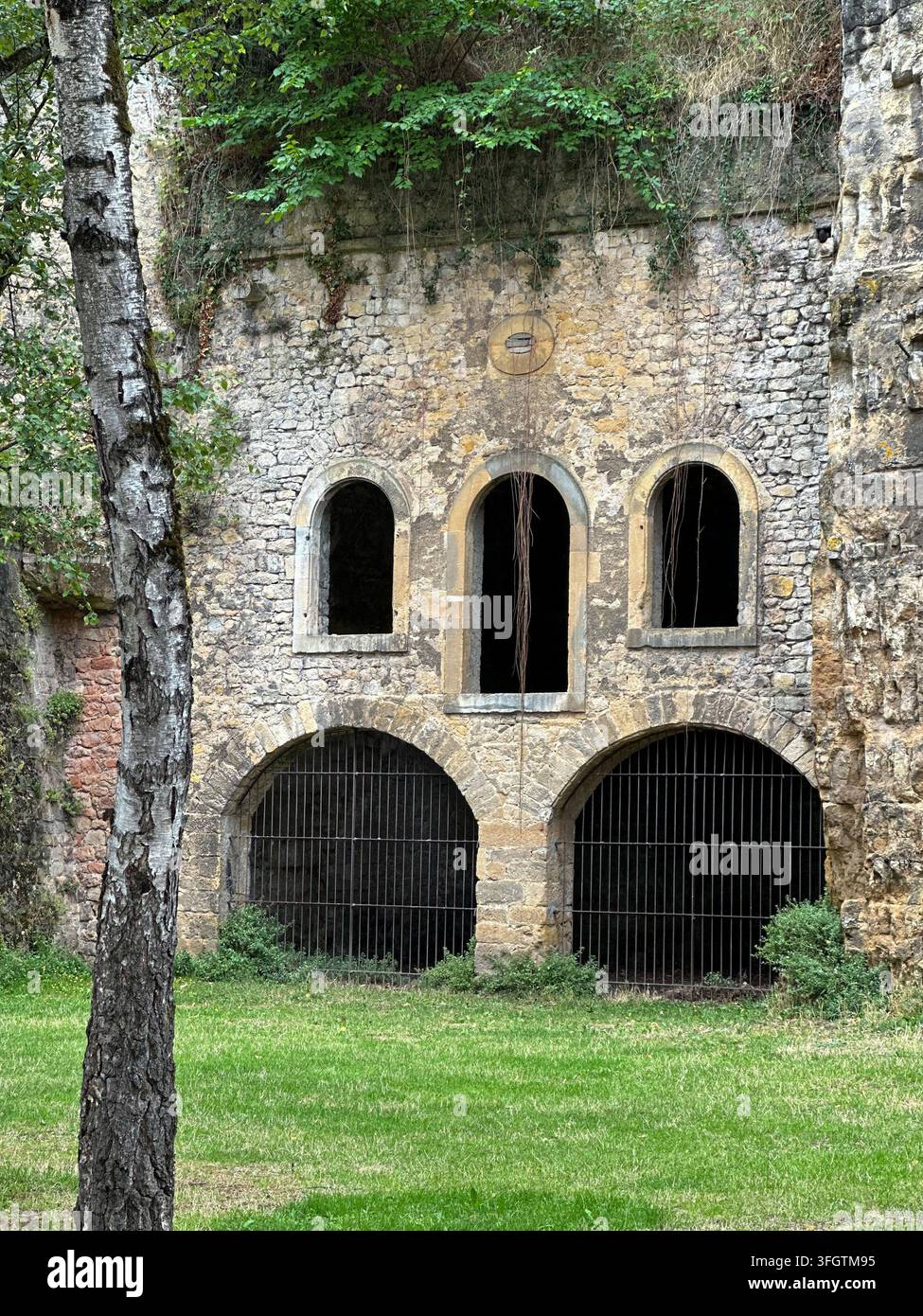 Alte Steinmauer einer Festung mit gewölbten Fenstern und vergitterten Tunneldurchgängen. Verwitterte Steinmauer einer Festung mit drei dunklen gewölbten oberen Fenstern a Stockfoto