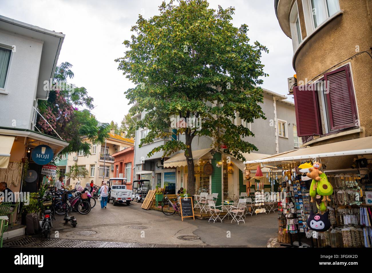Adalar, Türkei - 28. August 2026: Straßenansicht eines charmanten Marktbereichs mit einem großen Baum, Geschäften, Fahrrädern und Außensitzgelegenheiten in einem Wohngebiet Stockfoto