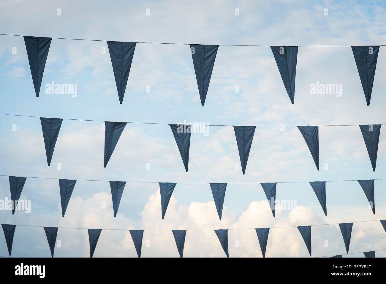 Dreieckige marineblaue Wimpelgirlande, die vor einem hellblauen Himmel mit sanften weißen Wolken hängt und ein klares grafisches Muster aus sich wiederholenden Flaggen erzeugt. Stockfoto