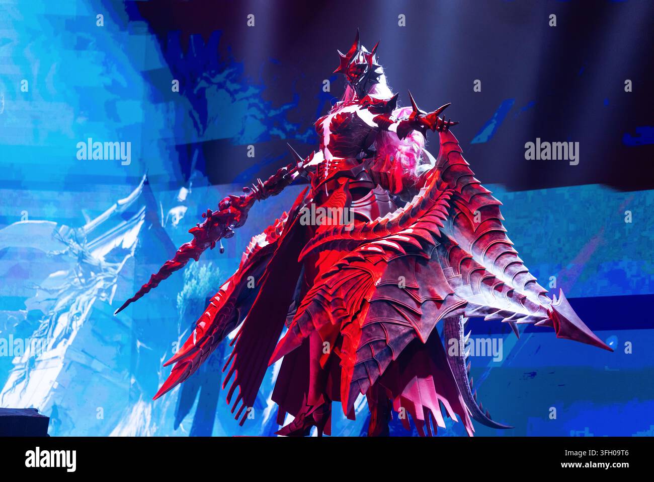 Köln, Deutschland – 26. August 2026: Fantasy-Charakter in auffälliger roter Rüstung mit Stacheln und einer Waffe, präsentiert auf einer Spielemesse mit farbenfrohem Licht Stockfoto