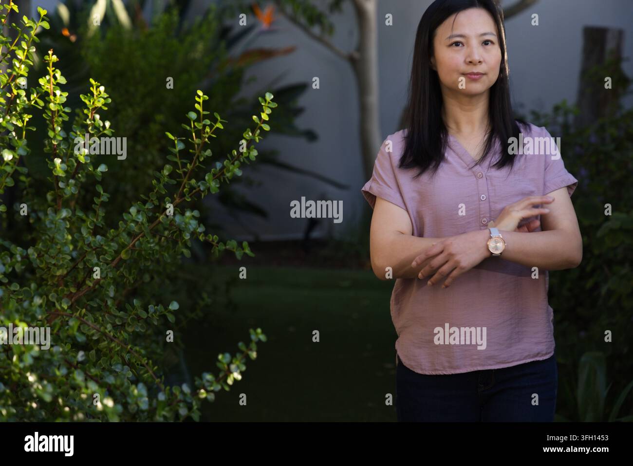 Reife asiatische Frau steht im Hinterhofgarten, passt ihre Armbanduhr an und trägt eine malvenfarbene Bluse Stockfoto