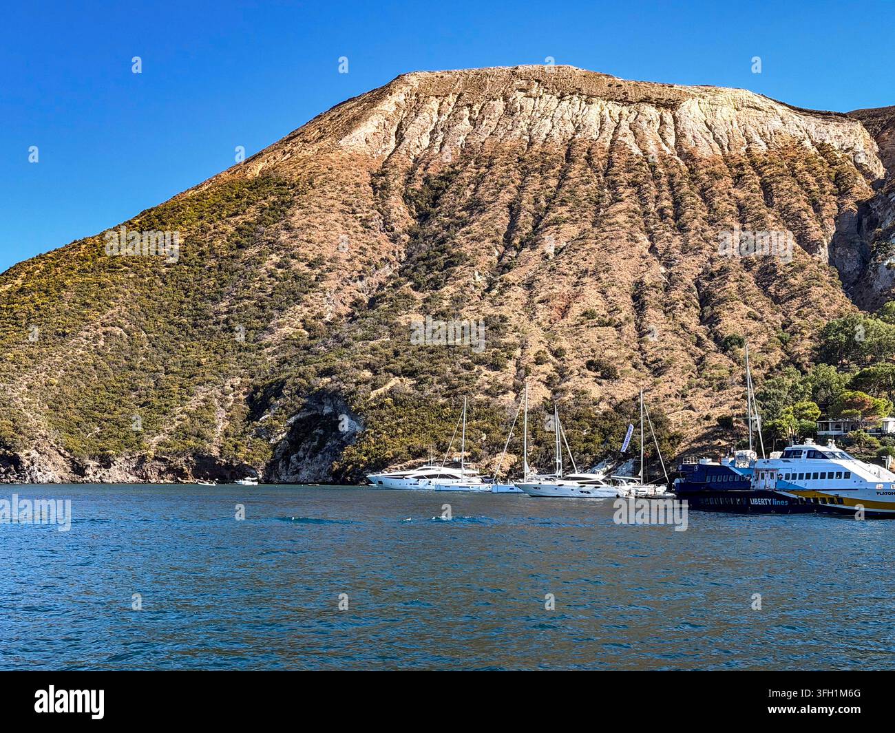 Vulcano, ME - IT - 5. Sep 2026: Steiler vulkanischer Hang erhebt sich über dem Hafen auf der Insel Vulcano, wo Segelboote, Yachten und Passagierfähren liegen Stockfoto