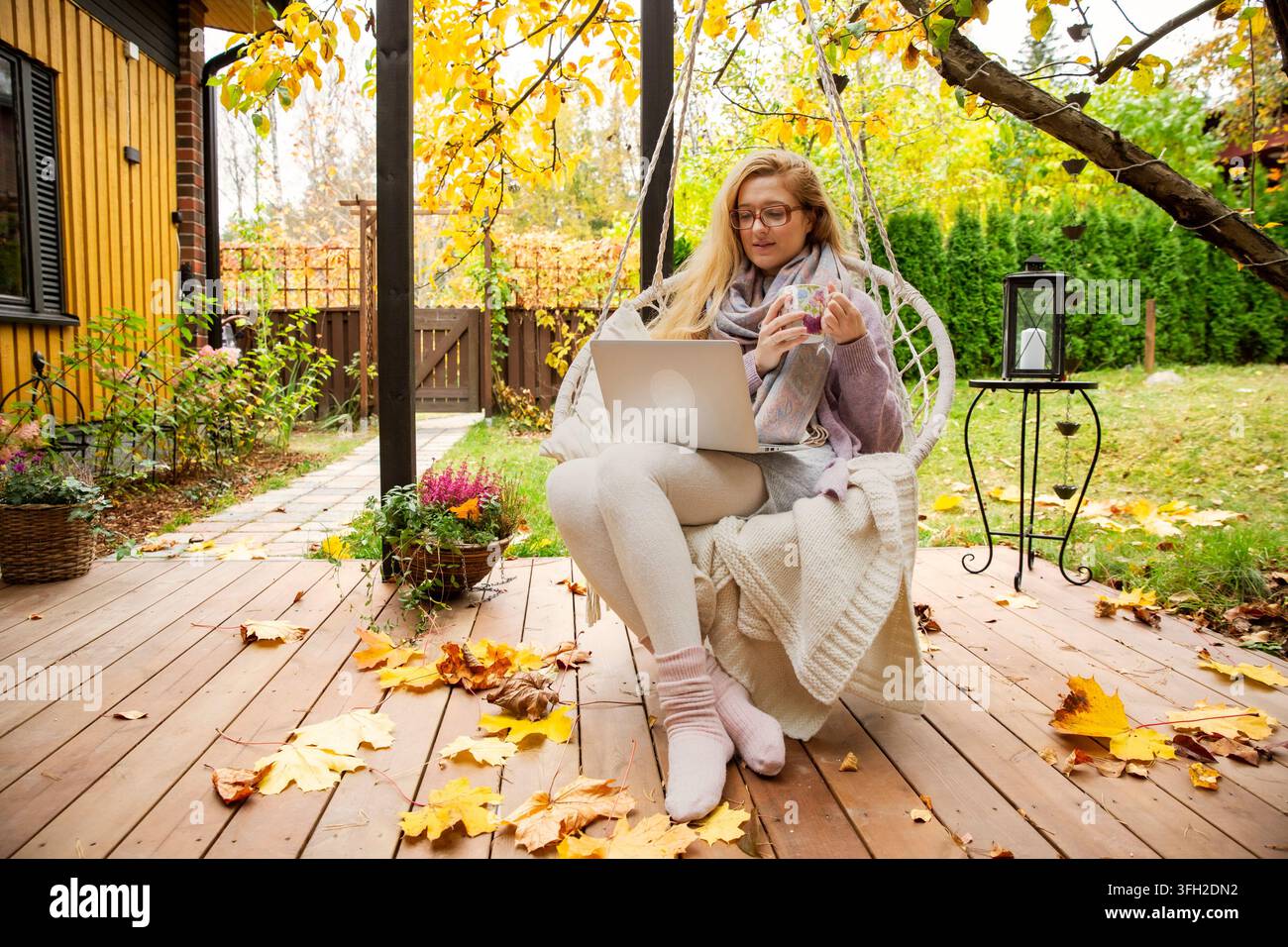 Eine mittelalte Frau arbeitet remote an einem Laptop, während sie Tee trinkt, in einem Hängesessel auf einer Herbstterrasse. Stockfoto