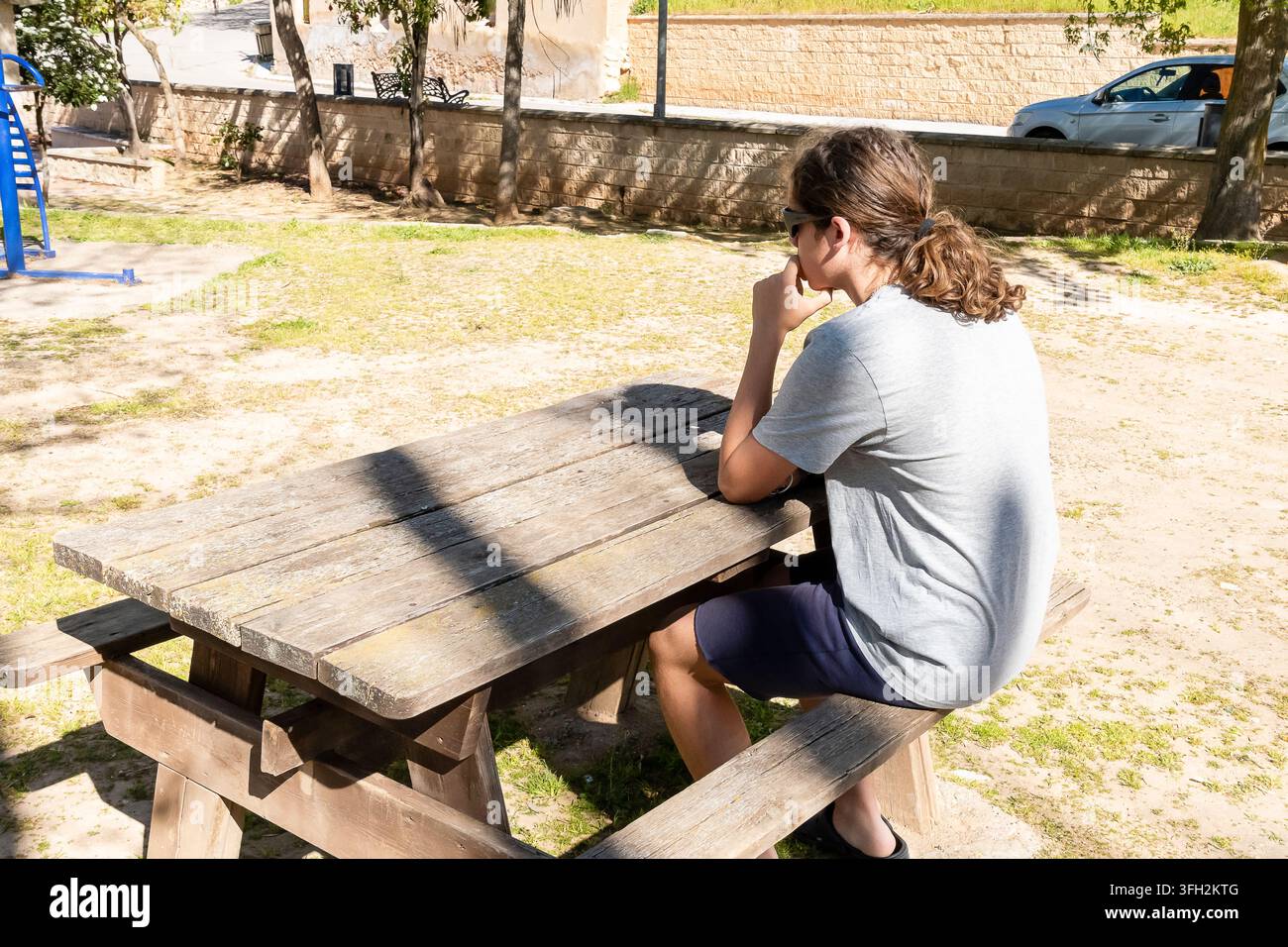 Eine Person sitzt an einem hölzernen Picknicktisch in einem Park, versunken in Gedanken, während sie das Sonnenlicht genießt. Stockfoto