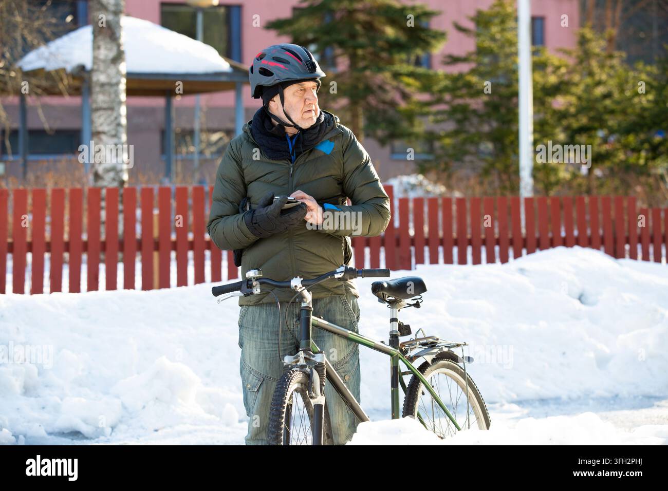 Ein älterer Mann überprüft sein Smartphone in der Nähe seines Fahrrads auf einer verschneiten Parkstraße in Helsinki, Finnland, und plant seine Winterpendelreise Stockfoto