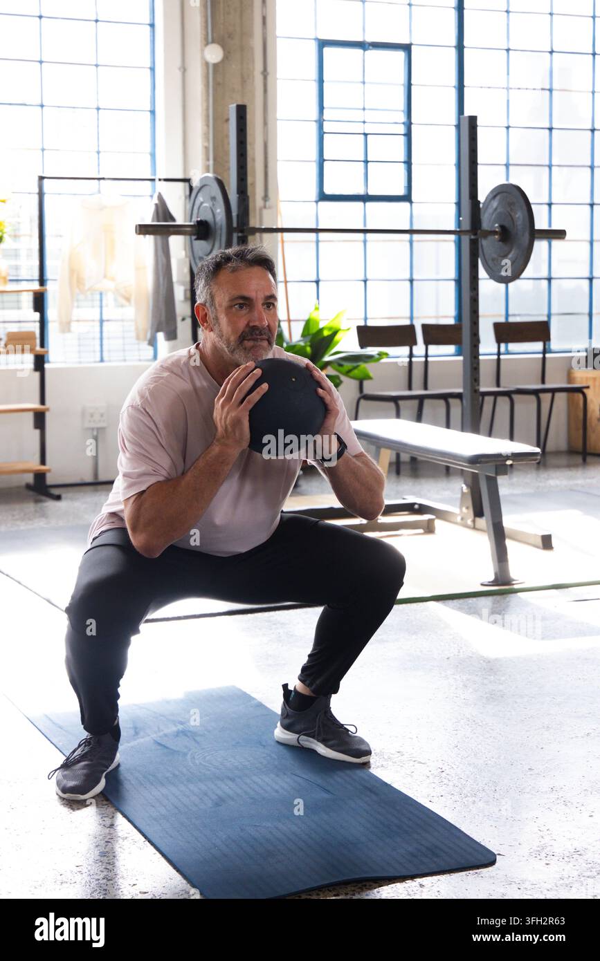 Mittealter Mann in der Hocke, der einen schwarzen Medizinball auf einer Matte in einem Fitnessstudio in der Nähe des Hantelständers hält Stockfoto