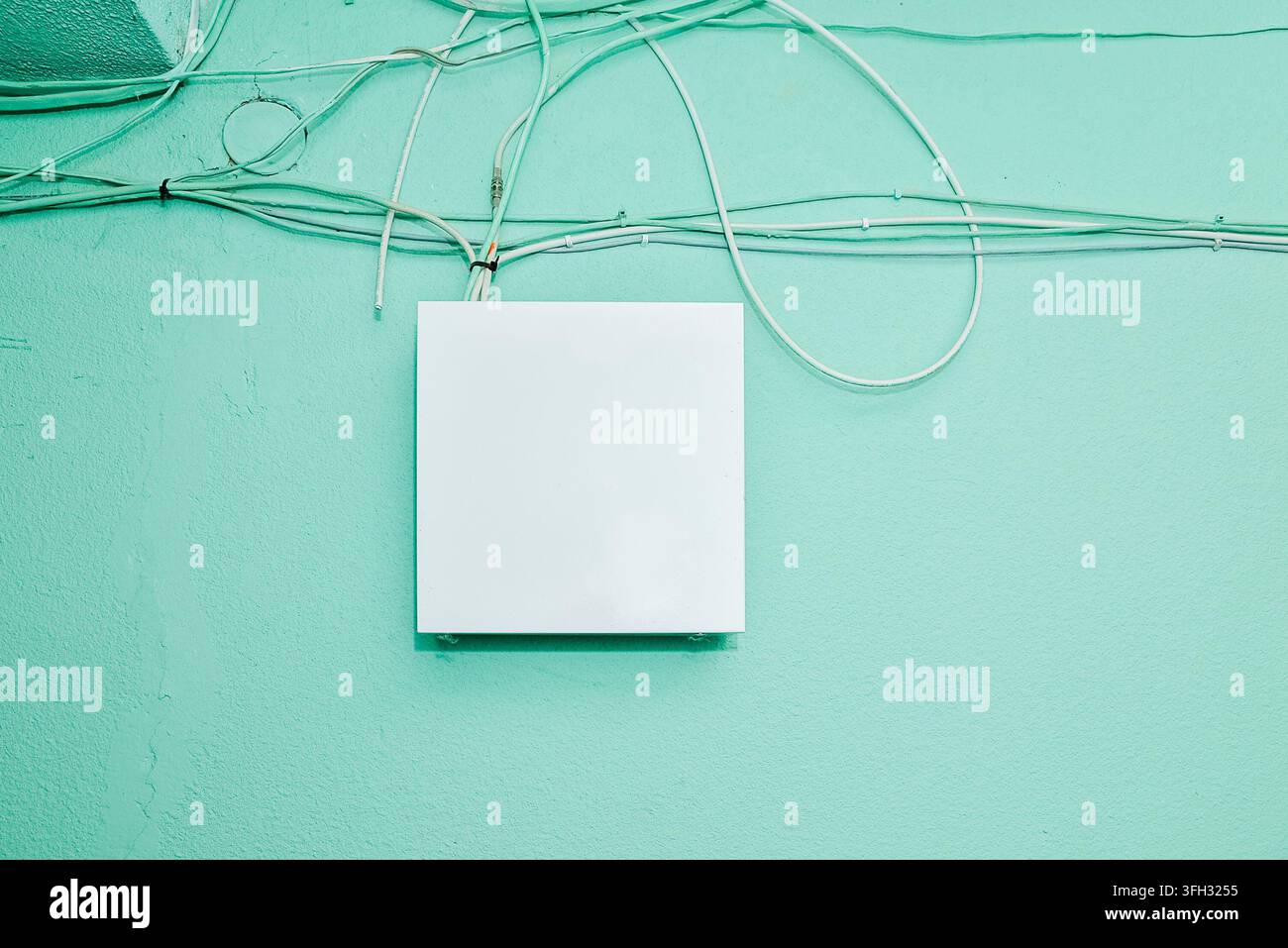 Weißes, leeres, rechteckiges Elektro- oder Anschlussdosenabdeckungs-Mockup-Panel an einer strukturierten mintgrünen Wand mit Kabeln. Stockfoto
