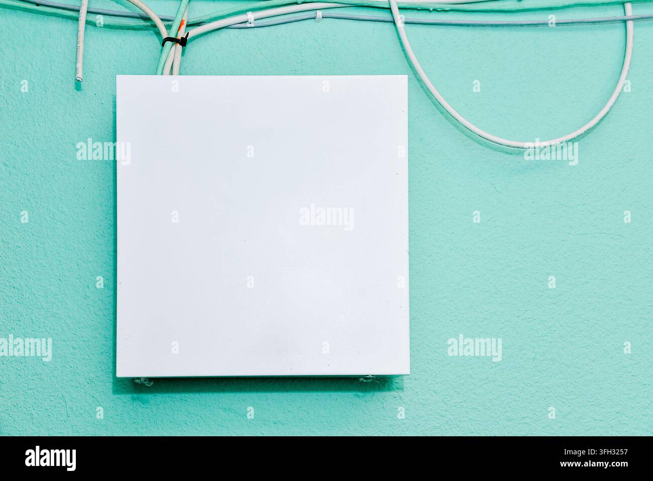 Weißes, leeres, rechteckiges Elektro- oder Anschlussdosenabdeckungs-Mockup-Panel an einer strukturierten mintgrünen Wand mit Kabeln. Stockfoto