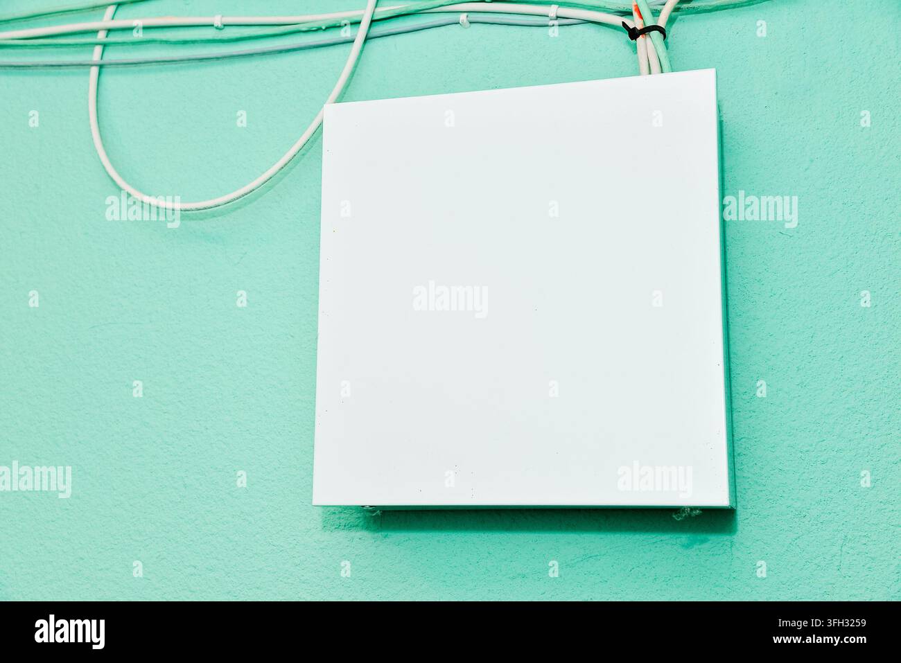 Weißes, leeres, rechteckiges Elektro- oder Anschlussdosenabdeckungs-Mockup-Panel an einer strukturierten mintgrünen Wand mit Kabeln. Stockfoto