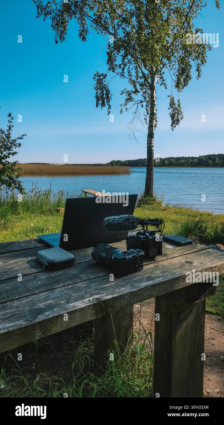 Laptop, Kamera und Mikrofon, die auf einem rustikalen Holztisch an einem ruhigen See angeordnet sind, repräsentieren einen flexiblen Arbeitsplatz für Digitalnomaden und Creator Stockfoto