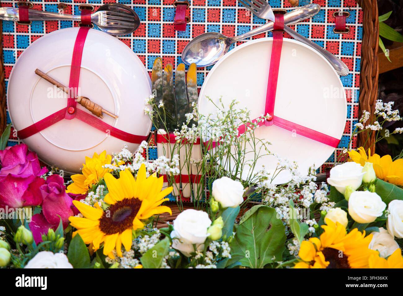 Dekorativer Tischgedeck mit weißen Tellern, silbernem Besteck, rosa Bandakzenten, bunten Blumen, gemusterter Kulisse und gewebten Korbdetails für Stockfoto