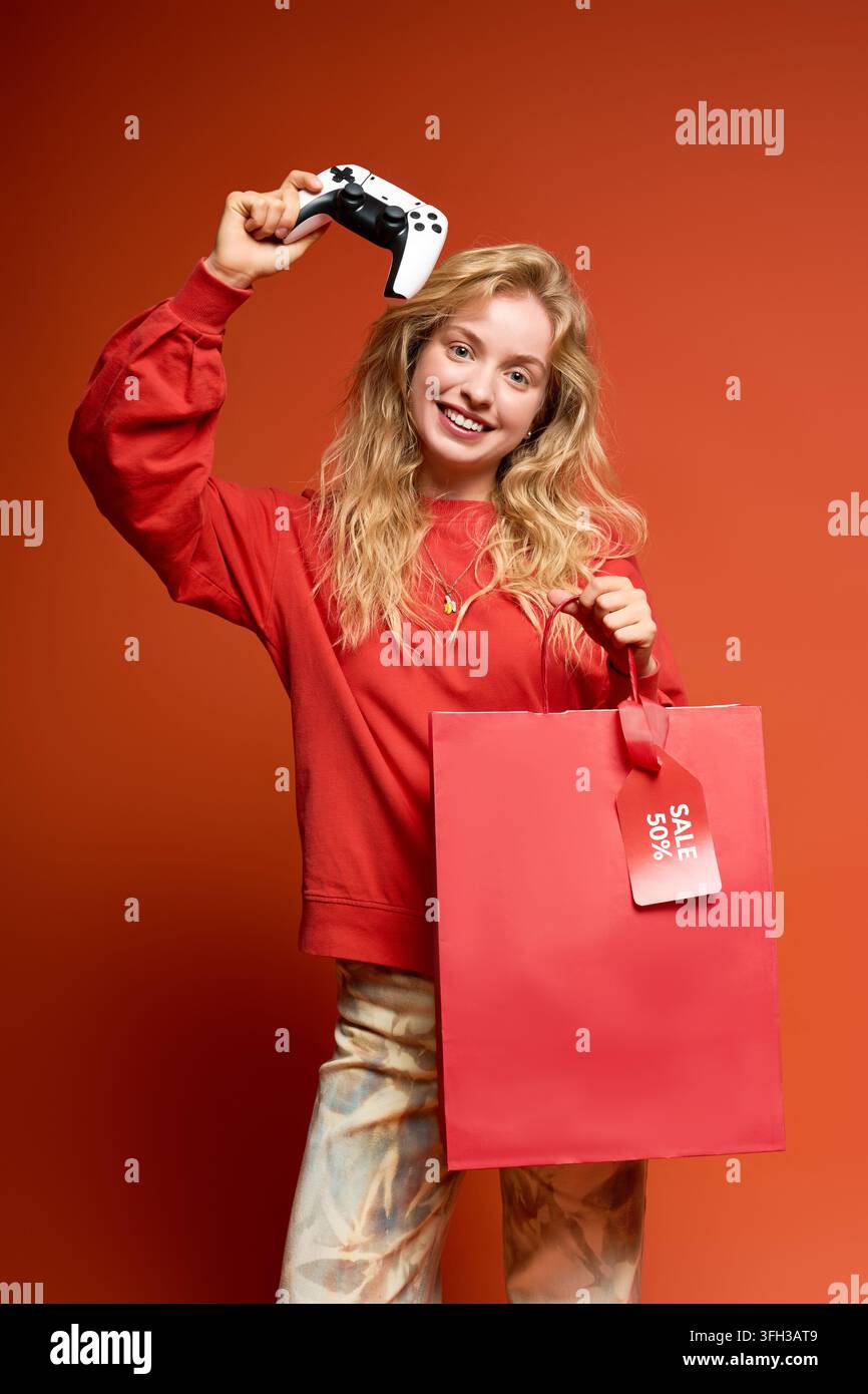 Junge blonde Frau hält freudig einen Game-Controller nach einem erfolgreichen Einkaufstag. Stockfoto