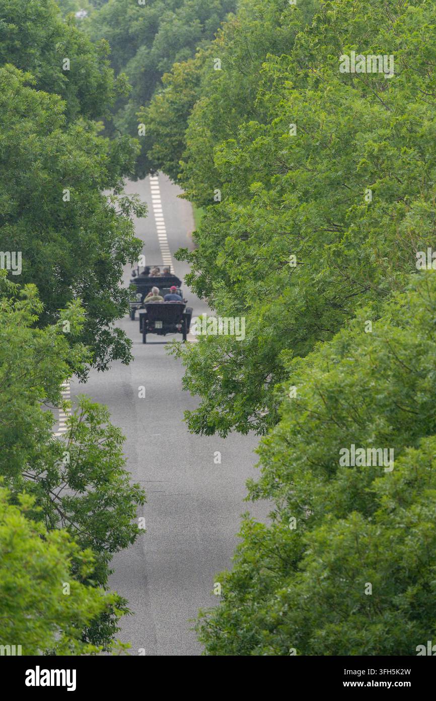 Eine Hochwinkel-Aufnahme zweier klassischer schwarzer Oldtimer, die eine graue Asphaltstraße entlangfahren, die von üppigen grünen Bäumen gesäumt ist, in einer malerischen Landschaft Stockfoto