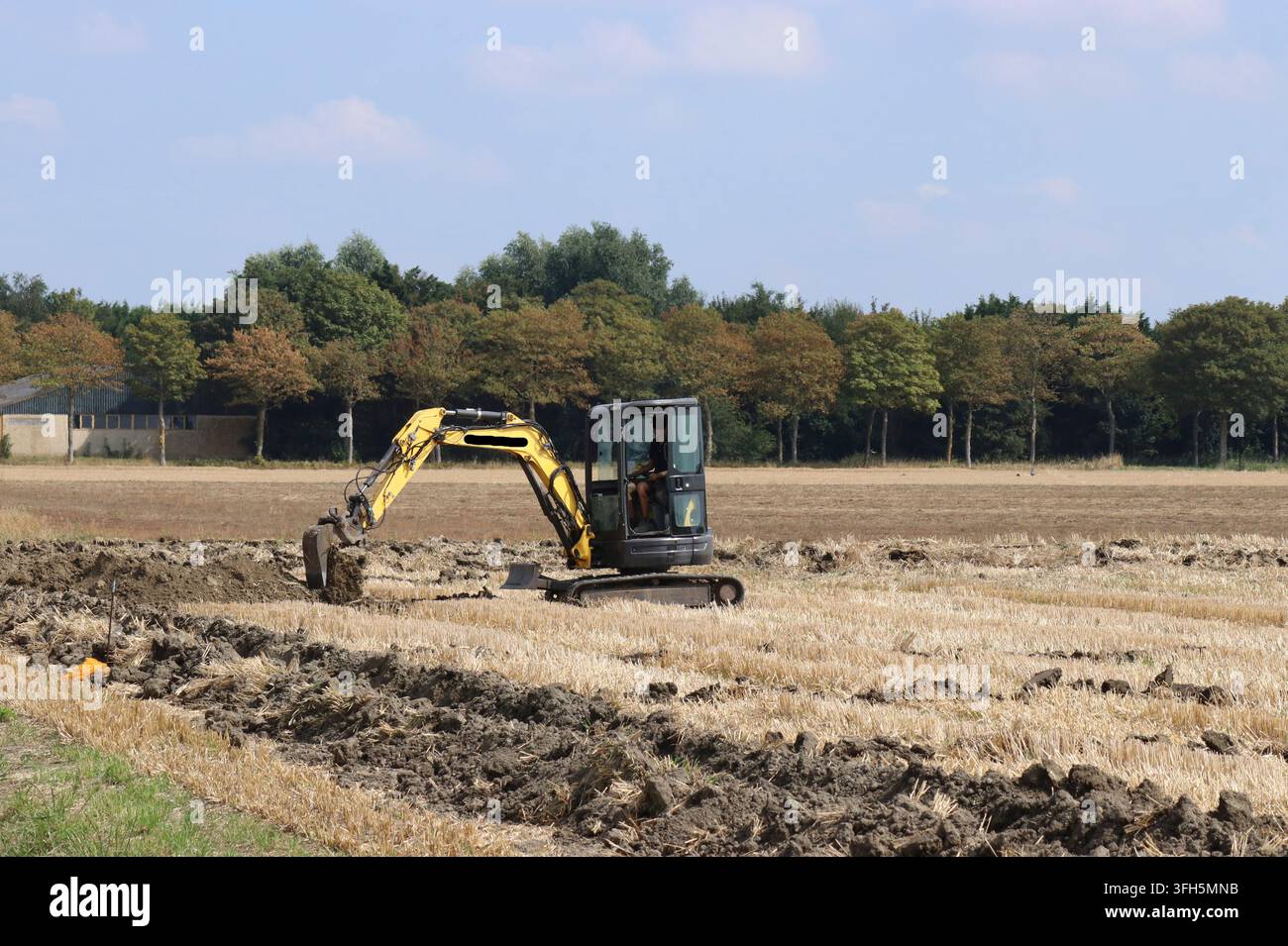 ein kleiner Bagger gräbt Gräben für die Installation von Abwasserrohren auf einem Feld im Sommer Stockfoto