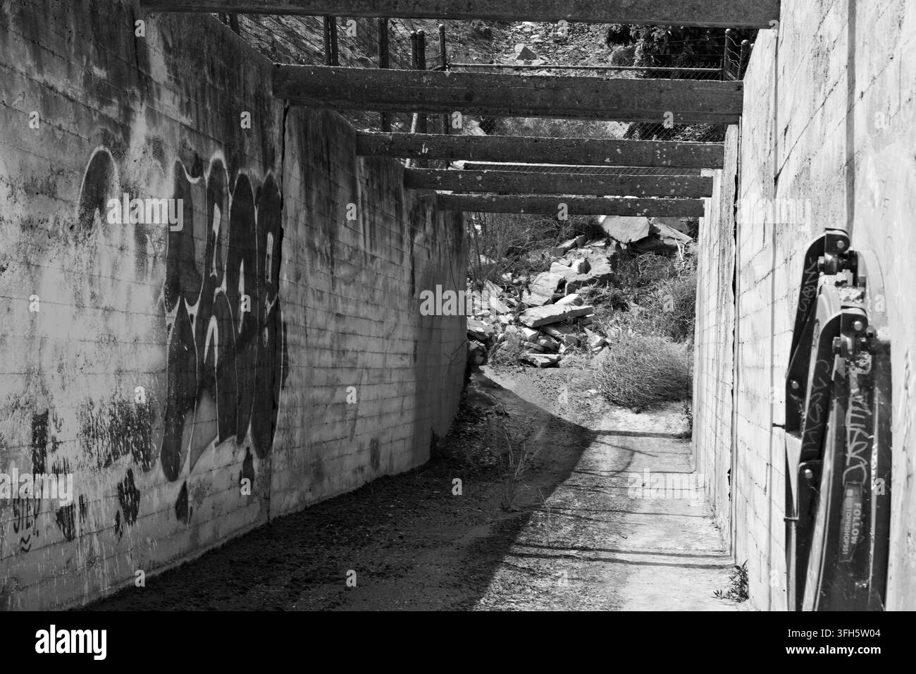 Verwitterte Betonwände und sich wiederholende Überkopfträger bilden einen schmalen trockenen Kanal mit tiefen Schatten und spärlichem Unkraut in Riverside, Kalifornien. Stockfoto