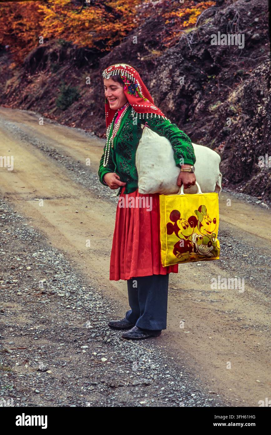 Junge Frau in farbenfrohem traditionellem Volkskostüm, die ein Bündel und eine Einkaufstasche entlang einer Schotterstraße trägt. Librazhd, Qark Elbasan, Albanien, Europa Stockfoto