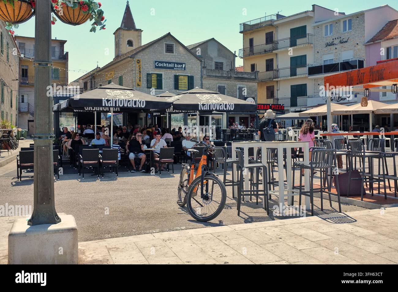 Pirovac, Kroatien, 16. August 2026: Belebter Uferplatz mit Außengastronomie, geparktem Fahrrad, schattigen Tischen, pastellfarbenen Fassaden und Kirchturm, der aufsteigt Stockfoto
