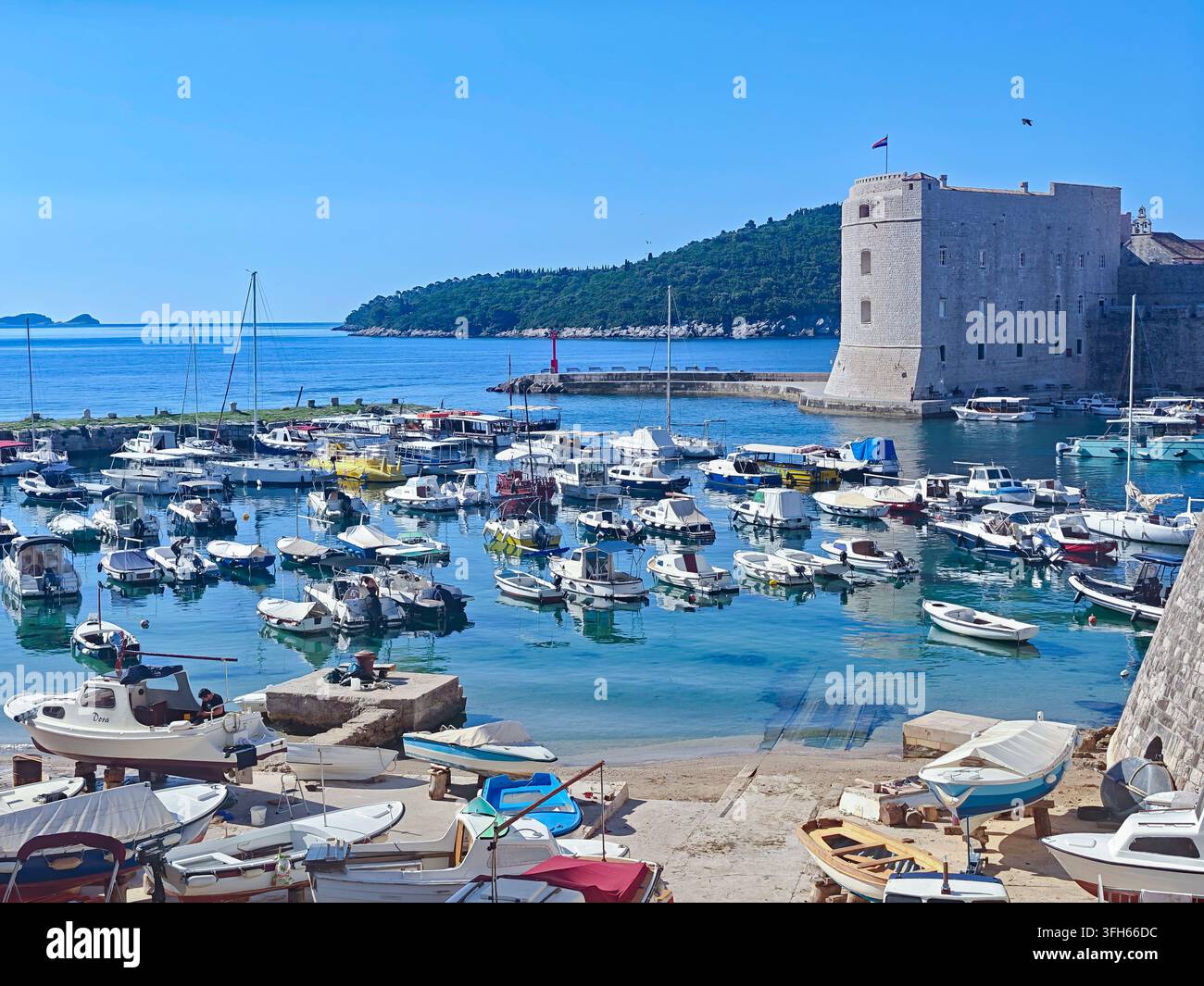 Zahlreiche kleine Boote, die in einem klaren blauen Meer in der Nähe einer alten Steinfestung unter einem strahlend sonnigen Himmel vor Anker liegen. - Smartphone-aufgenommenes Stockfoto