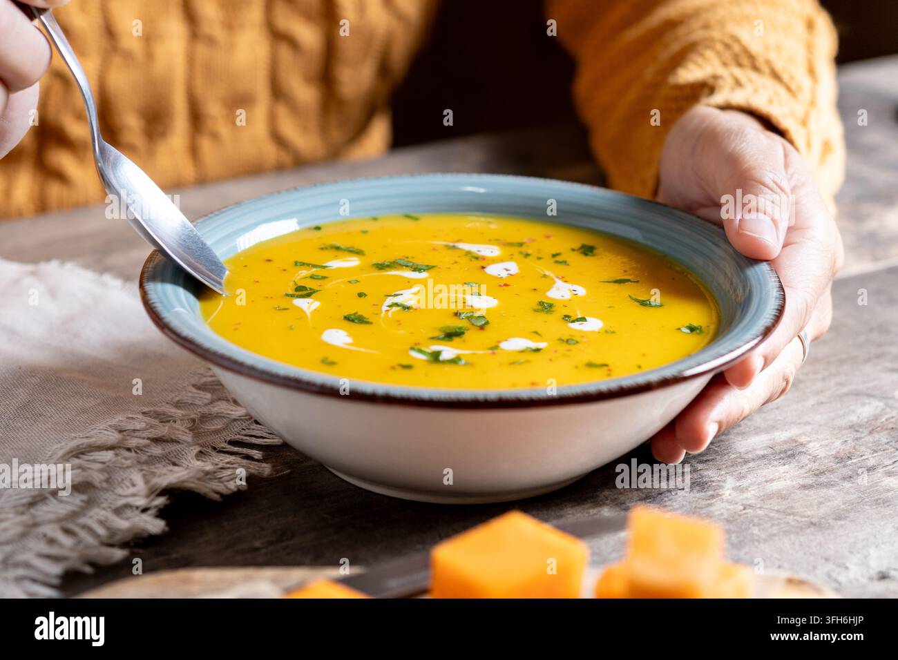 Nahaufnahme von Händen, die eine Schüssel mit cremiger Kürbissuppe halten, die mit frischen Kräutern und Sahne garniert ist, serviert mit einem Löffel auf einem rustikalen Tisch und gewürfeltem Kürbis-Kuchen Stockfoto