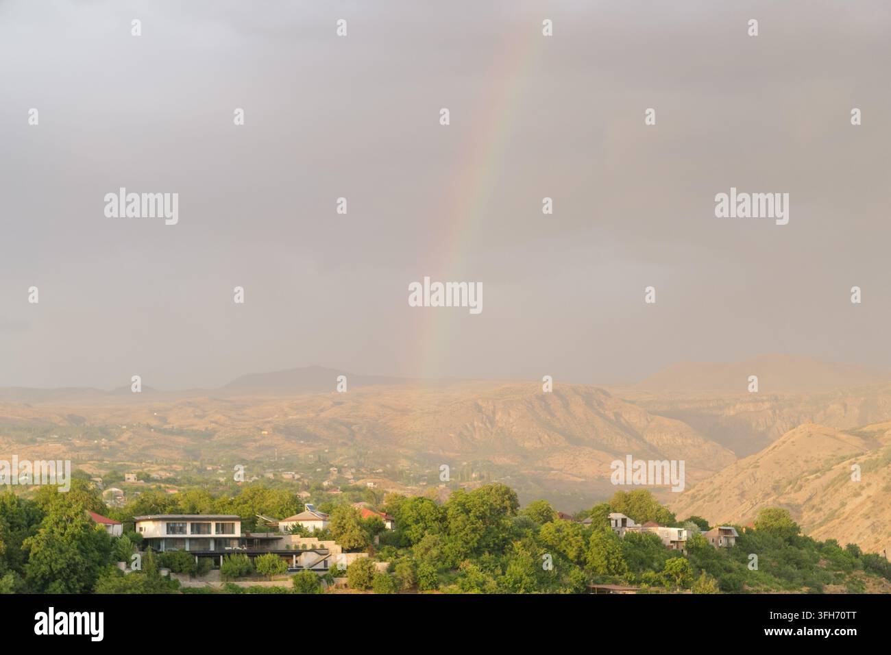 Blick auf sanfte Hügel und einen Regenbogen nach dem Regen, mit Häusern, die sich in üppiger Vegetation in einer bergigen Landschaft schmiegen Stockfoto